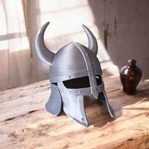 May include: A silver-colored Viking helmet with curved horns, rivets, and a face guard. The helmet sits on a wooden surface, with a dark brown bottle and a small glass holder with a brush in the background.