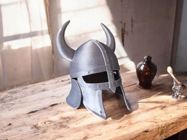May include: A silver-colored Viking helmet with curved horns, rivets, and a face guard. The helmet sits on a wooden surface, with a dark brown bottle and a small glass holder with a brush in the background.