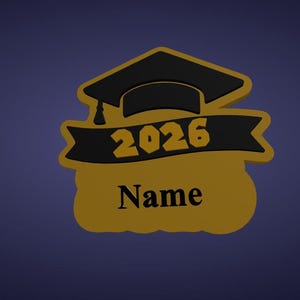 K&ouml;nnte beinhalten: Ein gold-schwarzes Abschluss-Emblem mit einem Mortarboard und Banner mit der Jahreszahl "2026" und dem Wort "Name". Das Design befindet sich vor einem dunkelblauen Hintergrund, ideal f&uuml;r personalisierte Abschlussgeschenke.