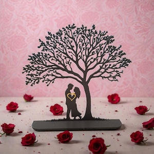 May include: A black metal tree sculpture with a couple embracing under its branches, a gold heart in the center. Red roses are scattered around the base. The sculpture sits on a gray base against a pink background.