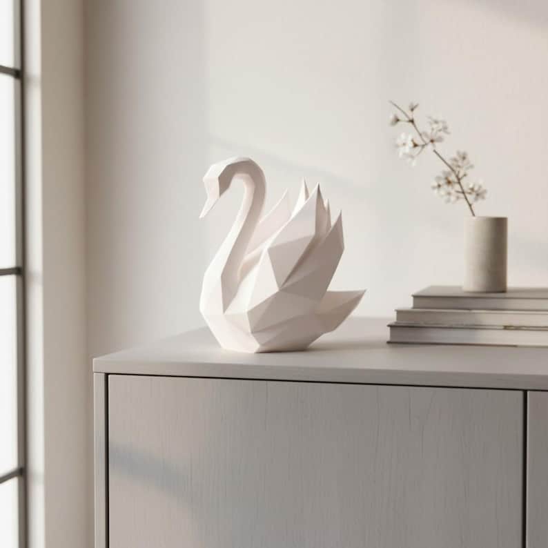May include: A white geometric swan sculpture with a modern, angular design. The swan is positioned on a light gray cabinet, with a window and a small vase of flowers in the background. The sculpture is a decorative home accent.