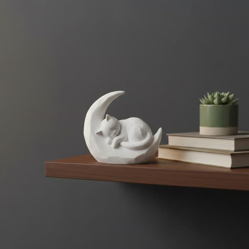Peut inclure: Figurine blanche repr&eacute;sentant un chat dormant sur un croissant de lune. Le chat est enroul&eacute; et la lune a une surface textur&eacute;e. La figurine est expos&eacute;e sur une &eacute;tag&egrave;re marron avec une petite plante en pot et des livres.