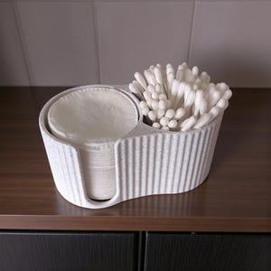 May include: A two-sectioned, light gray container with a textured, ribbed design. One side holds a stack of white cotton pads, and the other is filled with white cotton swabs. The container sits on a dark brown surface.