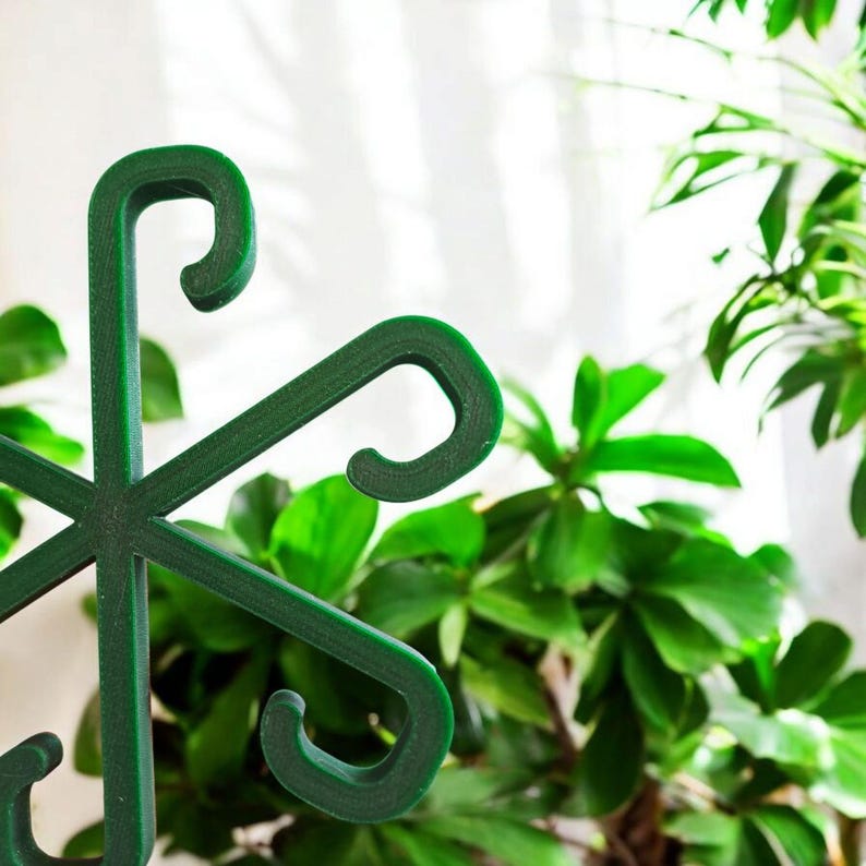 May include: Close-up of a dark green plant support structure. The support has a star-like design with curved hooks at the ends of each arm. The background features lush green plants and a white curtain.
