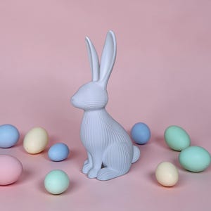 May include: A light blue ceramic bunny figurine with long ears and textured details, surrounded by pastel-colored Easter eggs. The bunny is sitting upright on a pink background, creating a festive spring scene.