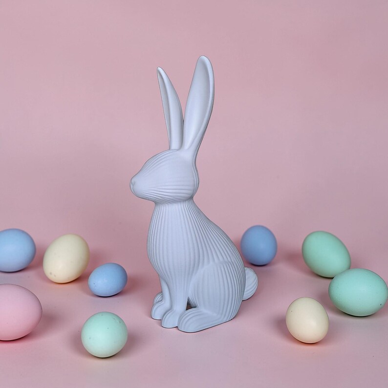 May include: A light blue ceramic bunny figurine with long ears and textured details, surrounded by pastel-colored Easter eggs. The bunny is sitting upright on a pink background, creating a festive spring scene.