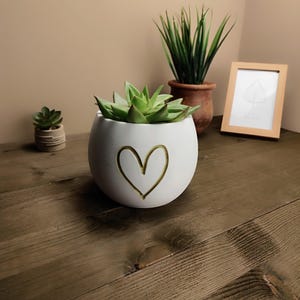Op de afbeelding: Een witte, ronde keramische plantenbak met een gouden hartontwerp, met een groene vetplant. Andere planten en een ingelijste foto staan op de achtergrond, alles op een houten oppervlak. Het thema is interieurdecoratie.