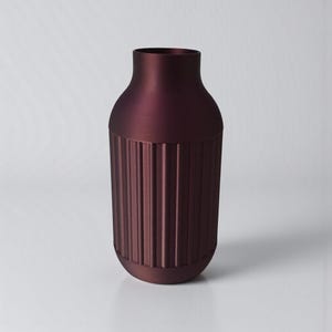May include: A burgundy-colored vase with a cylindrical lower section featuring vertical ribbed detailing. The upper portion of the vase has a smooth, rounded shape, and the vase has a matte finish. The vase is a decorative home decor item.