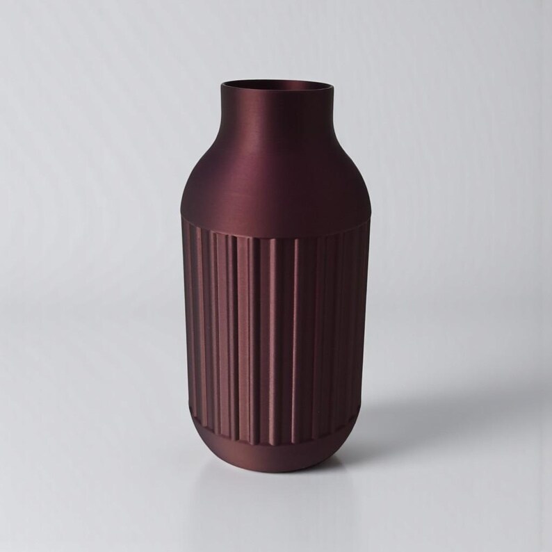 May include: A burgundy-colored vase with a cylindrical lower section featuring vertical ribbed detailing. The upper portion of the vase has a smooth, rounded shape, and the vase has a matte finish. The vase is a decorative home decor item.