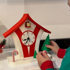 May include: A whimsical, red and white clock shaped like a house, with a white clock face and black hands. The clock has a red roof and red stripes. A person is holding a green Christmas tree and a candy cane. The clock is on a white surface.