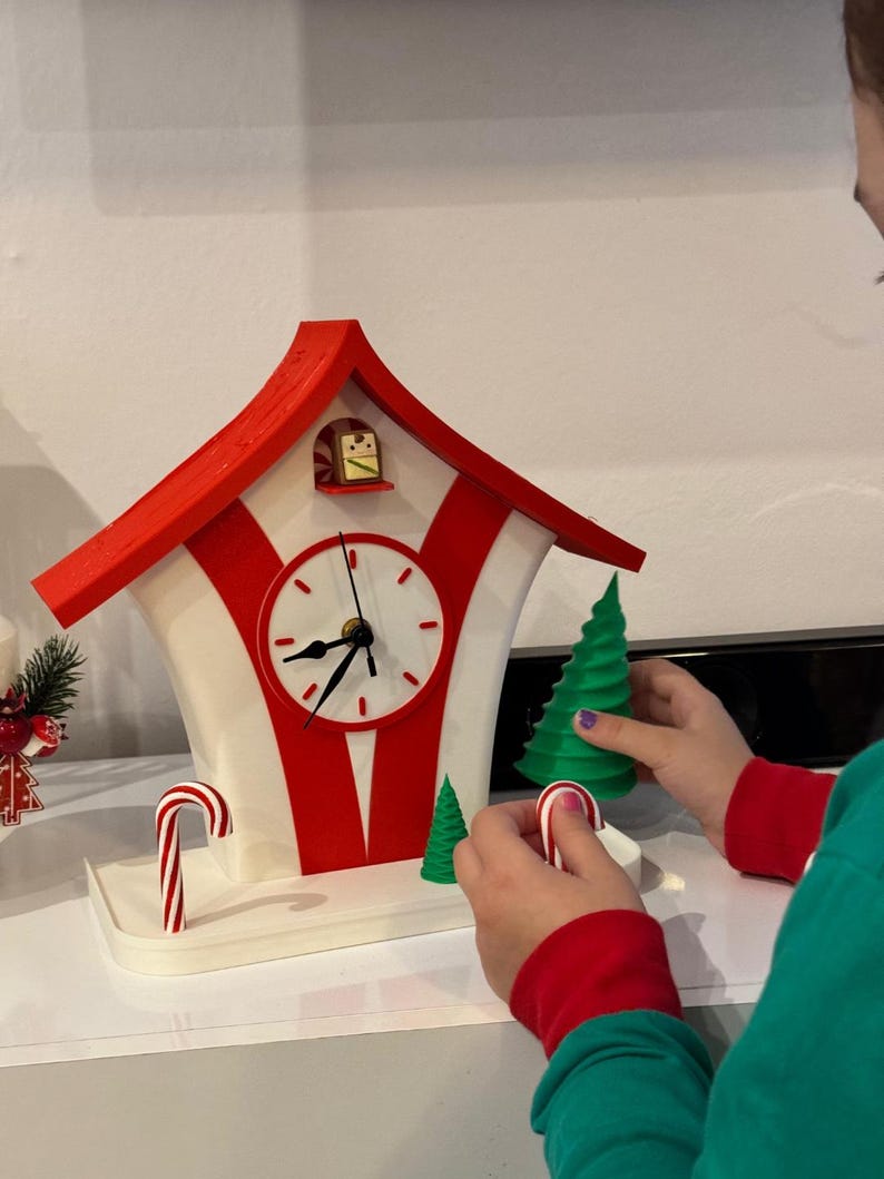 May include: A whimsical, red and white clock shaped like a house, with a white clock face and black hands. The clock has a red roof and red stripes. A person is holding a green Christmas tree and a candy cane. The clock is on a white surface.