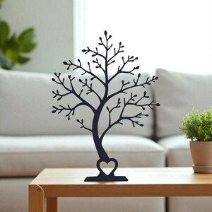 May include: A black metal tree-shaped jewelry holder with a heart-shaped cutout at the base. The tree has many branches and is displayed on a wooden table. A small potted plant is to the right.