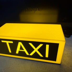 May include: A rectangular yellow taxi sign with the word "TAXI" in bold, white letters on a black background. The sign is illuminated, suggesting it is lit from within. The sign is a bright yellow color.