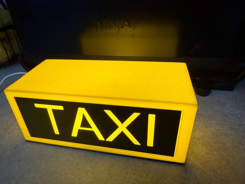 May include: A rectangular yellow taxi sign with the word "TAXI" in bold, white letters on a black background. The sign is illuminated, suggesting it is lit from within. The sign is a bright yellow color.