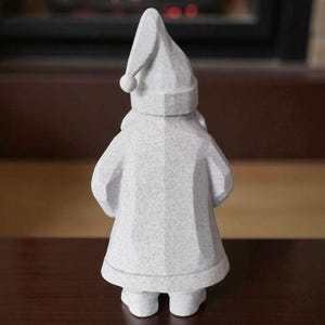 K&ouml;nnte beinhalten: Kleine, wei&szlig;e Zwergfigur mit spitzer M&uuml;tze und rundem Bommel. Der Zwerg tr&auml;gt einen langen Mantel und Stiefel. Die Figur besteht aus einem strukturierten Material und steht auf einer dunkelbraunen Oberfl&auml;che. Im Hintergrund ist ein Kamin zu sehen.