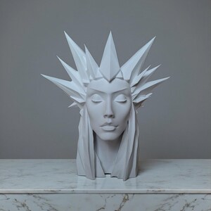 May include: A white sculpture of a woman's head with closed eyes and a geometric crown. The sculpture has long, flowing hair and is set against a neutral background. The bust is displayed on a marble-like surface.