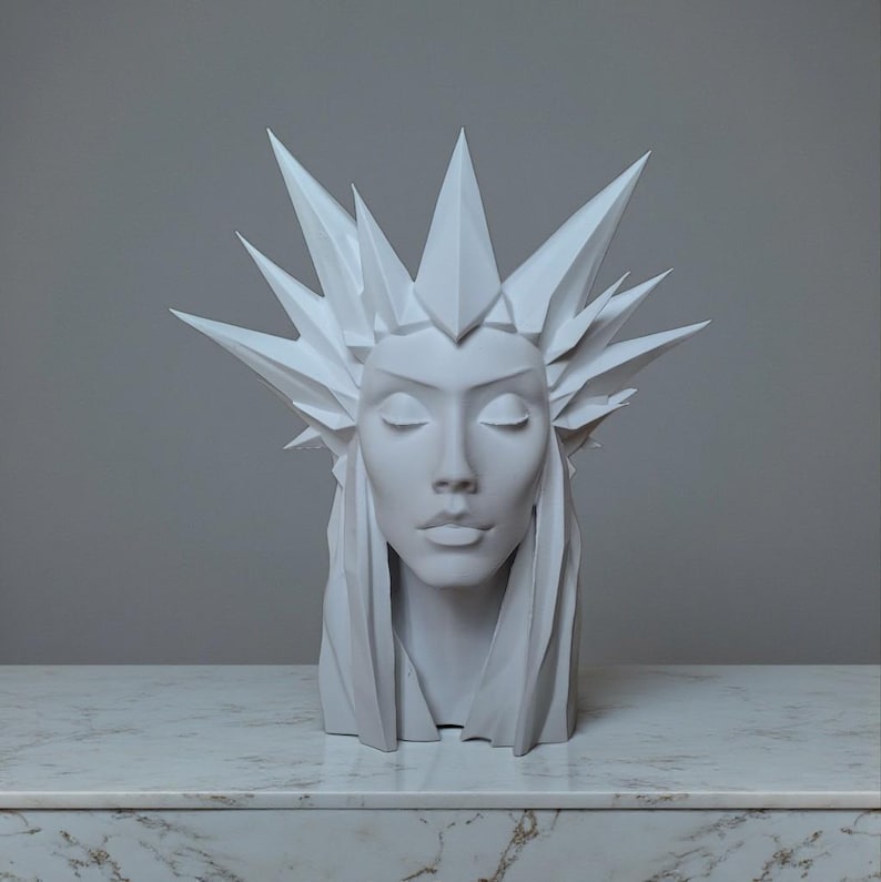 May include: A white sculpture of a woman's head with closed eyes and a geometric crown. The sculpture has long, flowing hair and is set against a neutral background. The bust is displayed on a marble-like surface.