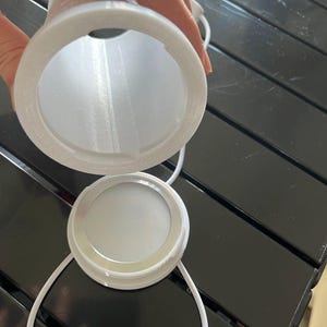 May include: A white, cylindrical object with a circular opening and a matching lid. The object appears to be made of a 3D-printed material. A white cord is attached to the lid. The object is held over a black surface.