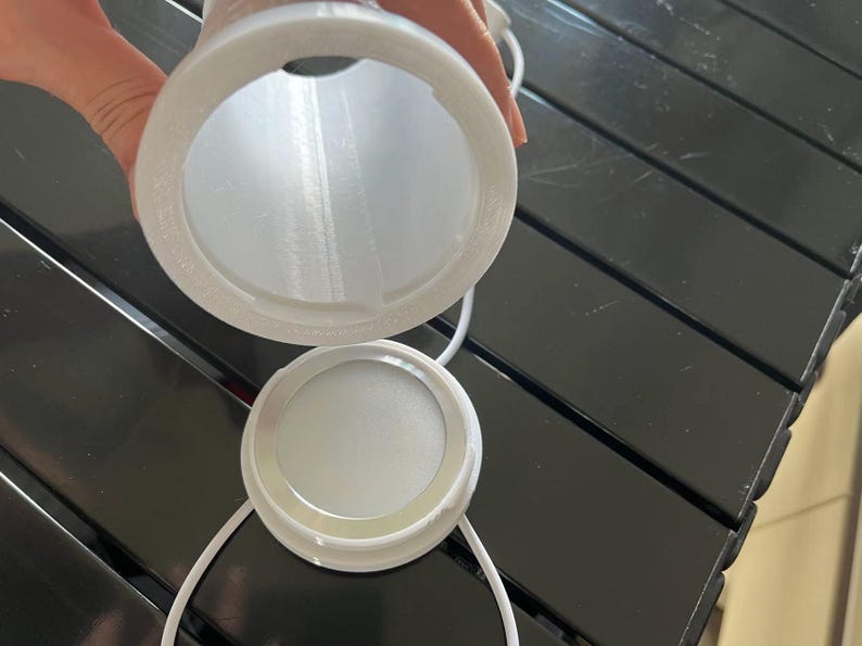 May include: A white, cylindrical object with a circular opening and a matching lid. The object appears to be made of a 3D-printed material. A white cord is attached to the lid. The object is held over a black surface.