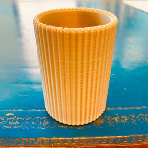 K&ouml;nnte beinhalten: Ein zylindrischer, hellbeige Becher mit vertikalen Rippendetails. Der Rand hat einen gewellten Rand. Der Becher besteht aus einem festen Material und steht auf einer blauen Oberfl&auml;che. Der Becher ist etwa 10 cm hoch.