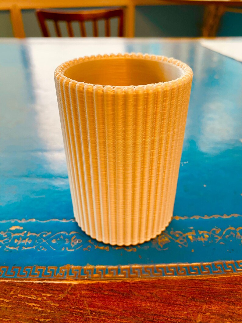 K&ouml;nnte beinhalten: Ein zylindrischer, hellbeige Becher mit vertikalen Rippendetails. Der Rand hat einen gewellten Rand. Der Becher besteht aus einem festen Material und steht auf einer blauen Oberfl&auml;che. Der Becher ist etwa 10 cm hoch.