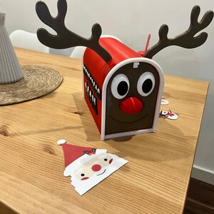 May include: A red and white Christmas mailbox shaped like a reindeer with large antlers, a red nose, and large eyes. A Santa Claus cutout is in the foreground. A vase with flowers is in the background.