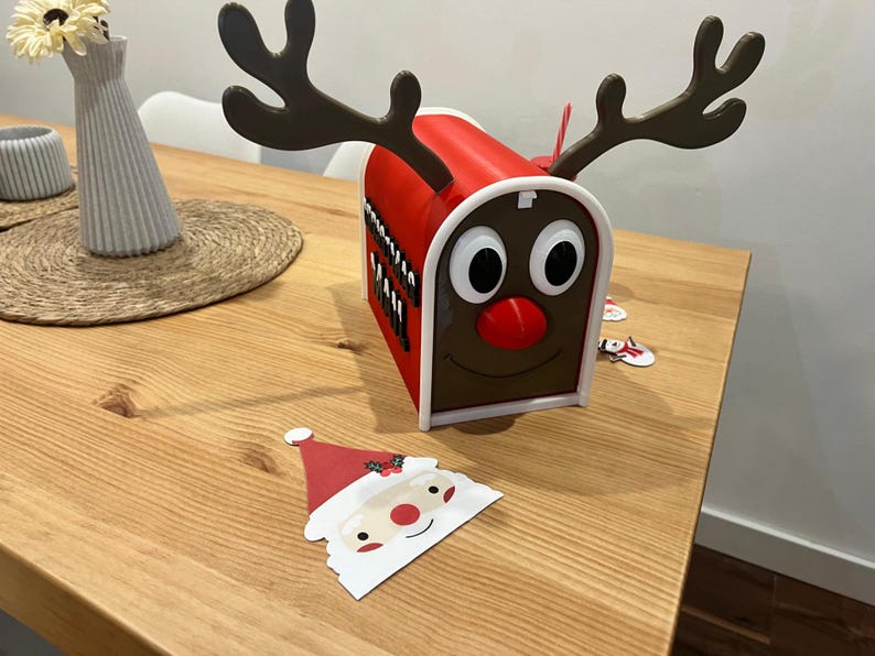 May include: A red and white Christmas mailbox shaped like a reindeer with large antlers, a red nose, and large eyes. A Santa Claus cutout is in the foreground. A vase with flowers is in the background.