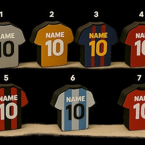 K&ouml;nnte beinhalten: Eine Sammlung von sieben Miniatur-Fu&szlig;balltrikots in verschiedenen Farben, jeweils mit der Nummer "10" und dem Wort "NAME" auf der R&uuml;ckseite. Die Trikots sind grau, gold, blau, rot, schwarz und hellblau.