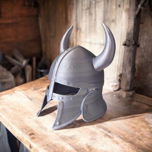 May include: A gray Viking helmet with curved horns, resting on a wooden surface. The helmet features a rounded crown, riveted details, and cheek guards. The background shows a rustic wooden interior.