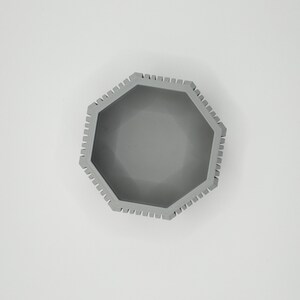 Puede incluir: Vista superior de un cuenco octogonal gris. El cuenco tiene un borde texturizado con peque&ntilde;as muescas espaciadas uniformemente. El interior es liso y del mismo color gris. El cuenco est&aacute; centrado sobre un fondo blanco.