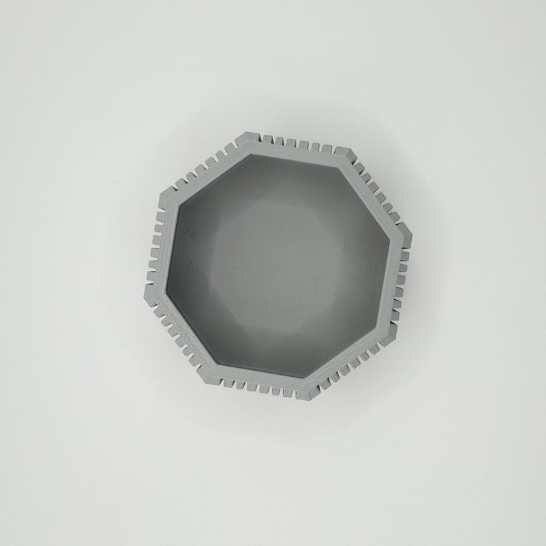 Puede incluir: Vista superior de un cuenco octogonal gris. El cuenco tiene un borde texturizado con peque&ntilde;as muescas espaciadas uniformemente. El interior es liso y del mismo color gris. El cuenco est&aacute; centrado sobre un fondo blanco.