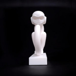 Puede incluir: Escultura blanca de un mono sentado, con los brazos alrededor de las piernas. La escultura est&aacute; sobre una base rectangular blanca, sobre un fondo negro. La cara del mono tiene ojos y orejas grandes y estilizados.