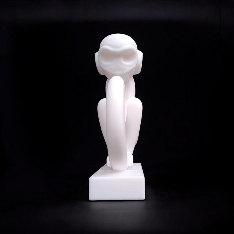 Puede incluir: Escultura blanca de un mono sentado, con los brazos alrededor de las piernas. La escultura est&aacute; sobre una base rectangular blanca, sobre un fondo negro. La cara del mono tiene ojos y orejas grandes y estilizados.