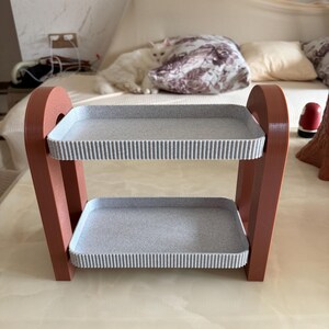 May include: A two-tiered decorative shelf with a modern design. The frame is a reddish-brown color, supporting two gray trays with a textured, ribbed edge. The shelf is designed to hold small items and is suitable for home decor.
