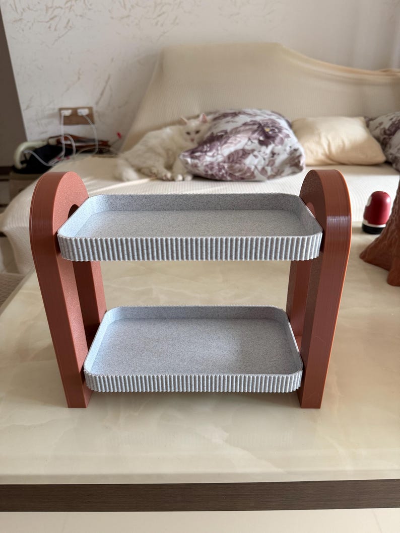 May include: A two-tiered decorative shelf with a modern design. The frame is a reddish-brown color, supporting two gray trays with a textured, ribbed edge. The shelf is designed to hold small items and is suitable for home decor.
