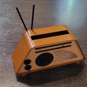 May include: A small, brown, retro-style radio with black antennas. The radio has a speaker, two knobs, and a tuning dial. The design is reminiscent of vintage radios from the mid-20th century.