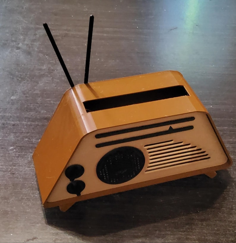 May include: A small, brown, retro-style radio with black antennas. The radio has a speaker, two knobs, and a tuning dial. The design is reminiscent of vintage radios from the mid-20th century.