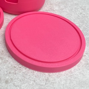 May include: A bright pink, round coaster with a raised edge. The coaster is made of a solid material and sits on a light gray surface. The coaster is part of a set, with a portion of another coaster and a cup visible.