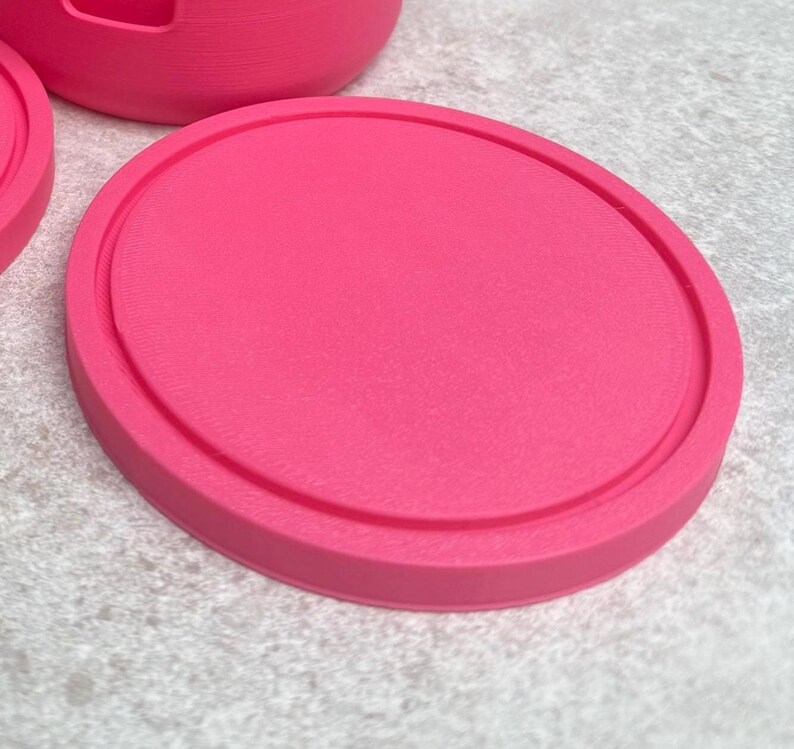 May include: A bright pink, round coaster with a raised edge. The coaster is made of a solid material and sits on a light gray surface. The coaster is part of a set, with a portion of another coaster and a cup visible.