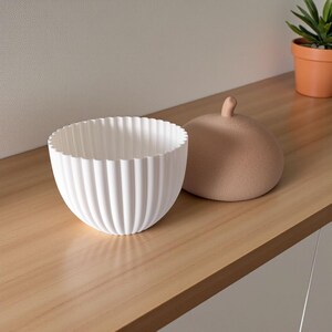 May include: A white, ribbed ceramic bowl with a scalloped rim sits next to a tan, rounded lid with a small knob. The bowl and lid are on a light wood surface, with a small potted plant in the background.