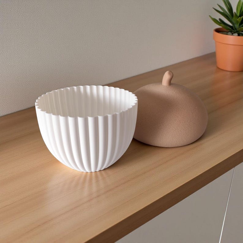 May include: A white, ribbed ceramic bowl with a scalloped rim sits next to a tan, rounded lid with a small knob. The bowl and lid are on a light wood surface, with a small potted plant in the background.