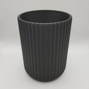 May include: A dark gray, cylindrical container with vertical ribbed detailing. The container has a wide opening and a rounded base. The surface has a matte finish. The container is suitable for various uses, such as a decorative vase or storage.