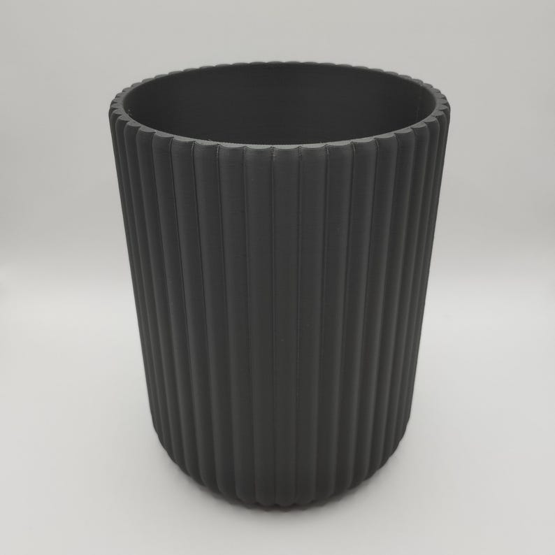 May include: A dark gray, cylindrical container with vertical ribbed detailing. The container has a wide opening and a rounded base. The surface has a matte finish. The container is suitable for various uses, such as a decorative vase or storage.