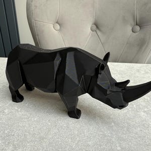 May include: A black geometric rhino sculpture. The rhino is made of multiple flat planes, giving it a modern, angular appearance. The sculpture is standing on a light gray surface.