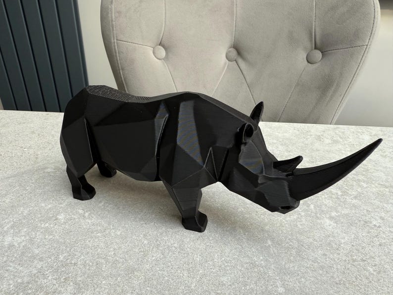 May include: A black geometric rhino sculpture. The rhino is made of multiple flat planes, giving it a modern, angular appearance. The sculpture is standing on a light gray surface.