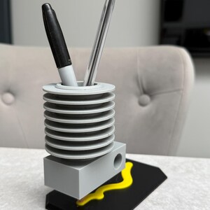 May include: A gray pen holder with a black base and yellow accent. The pen holder has a cylindrical design with a series of stacked, circular tiers. A black marker and a clear pen are inside the holder.