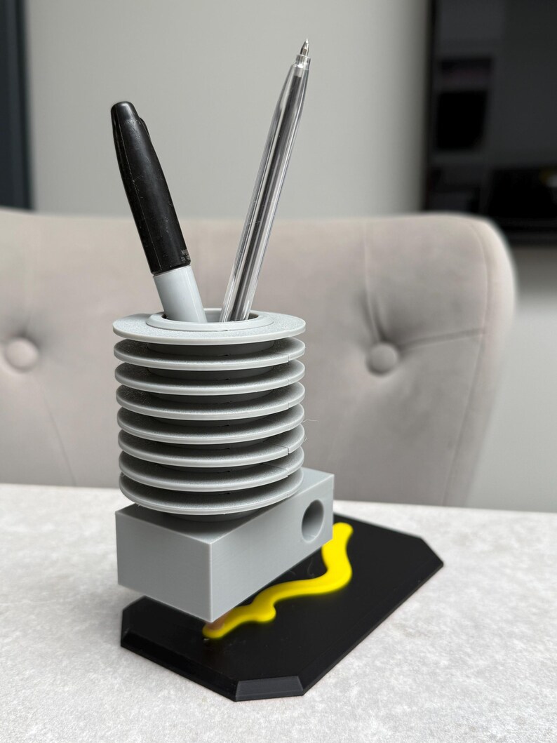 May include: A gray pen holder with a black base and yellow accent. The pen holder has a cylindrical design with a series of stacked, circular tiers. A black marker and a clear pen are inside the holder.