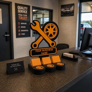 May include: An orange wrench and tire logo with the text "YOUR BUSINESS NAME" on a black base. The sign is on a counter with business card holders and a sign that says "THANK YOU FOR YOUR BUSINESS". A sign on the wall says "QUALITY SERVICE YOU CAN TRUST".