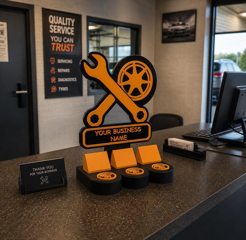 May include: An orange wrench and tire logo with the text "YOUR BUSINESS NAME" on a black base. The sign is on a counter with business card holders and a sign that says "THANK YOU FOR YOUR BUSINESS". A sign on the wall says "QUALITY SERVICE YOU CAN TRUST".