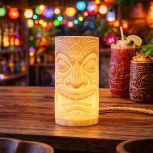 Peut inclure: Une lampe tiki cr&egrave;me avec un visage sculpt&eacute;, &eacute;clair&eacute;e de l'int&eacute;rieur. La lampe est pos&eacute;e sur un bar en bois &agrave; c&ocirc;t&eacute; de cocktails dans des verres d&eacute;coratifs. L'arri&egrave;re-plan pr&eacute;sente un bar avec des lumi&egrave;res color&eacute;es.